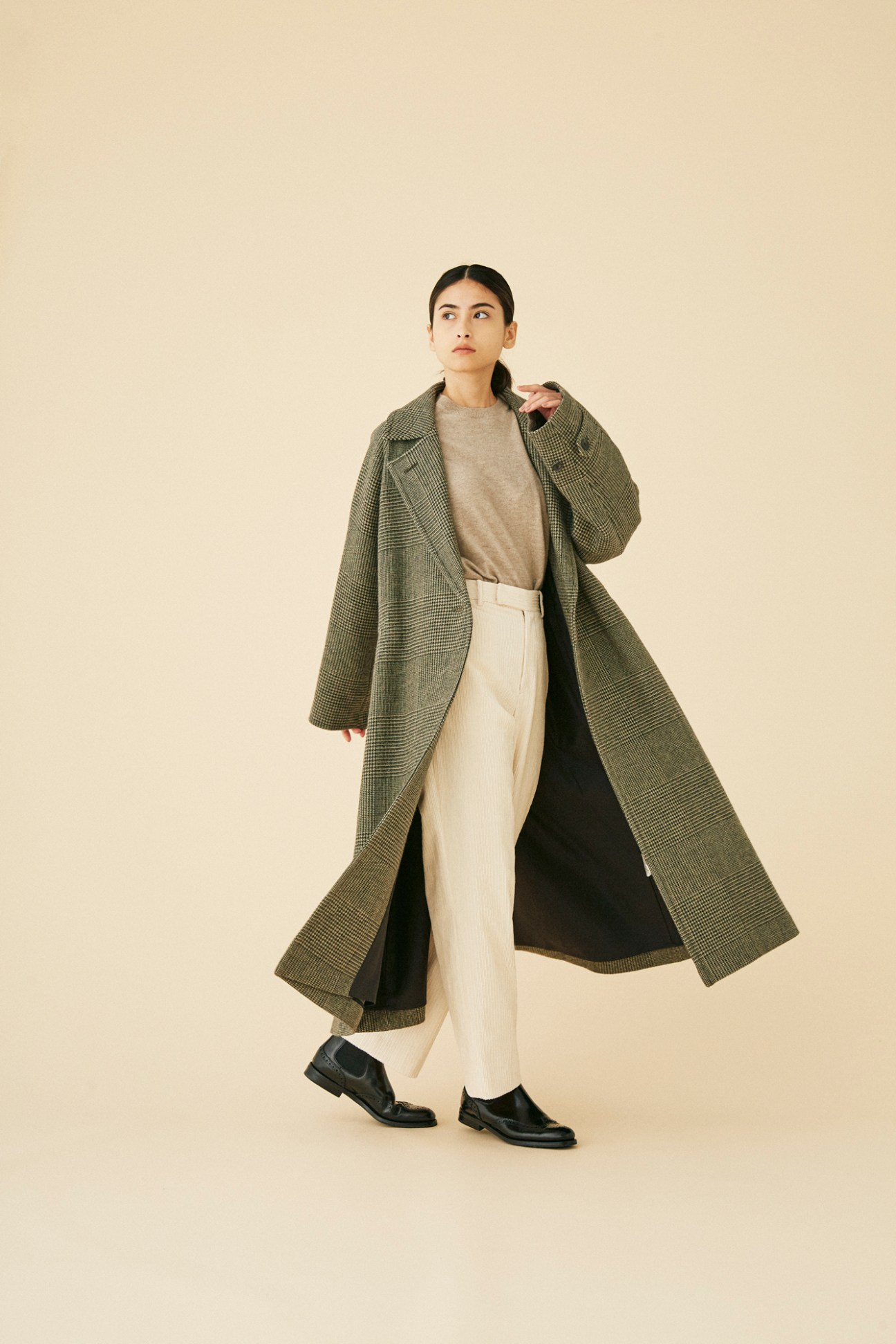 Woman wearing long olive green checkered coat by Yleve in minimalist studio setting