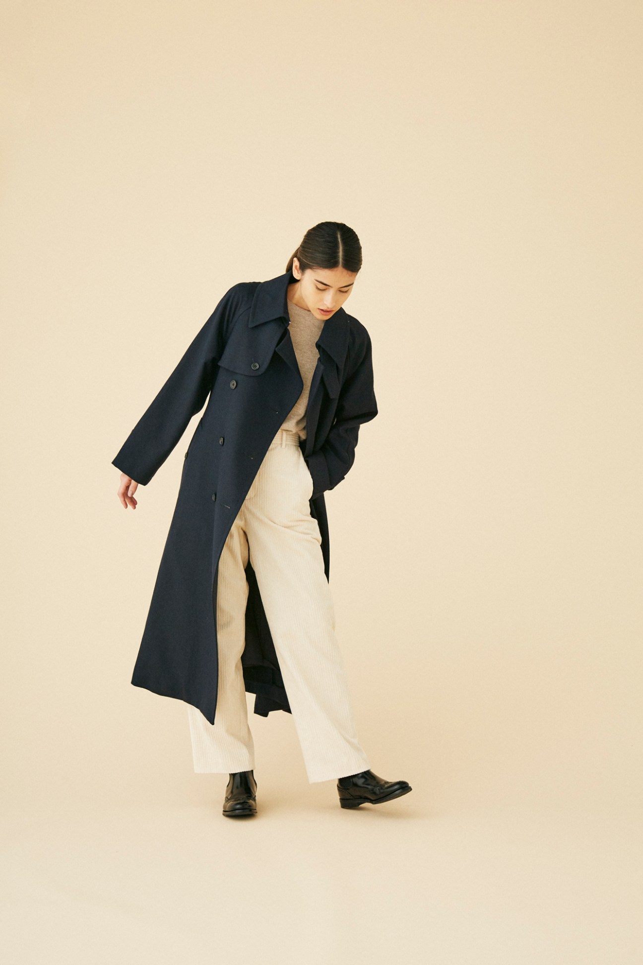 Woman wearing navy trench coat by Margaret Howell over beige sweater and cream trousers in studio setting