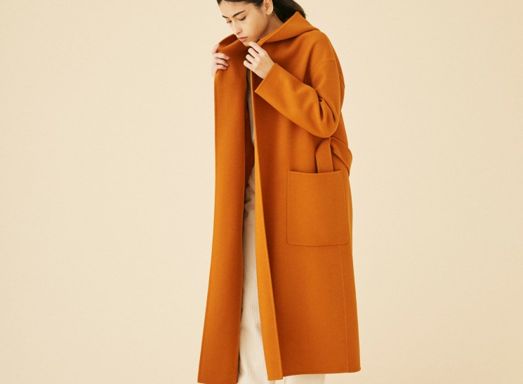 Woman wearing long orange wool coat by United Arrows against neutral studio background