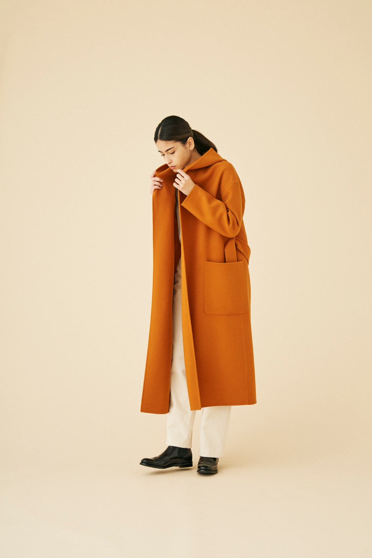 Woman wearing long orange wool coat by United Arrows against neutral studio background