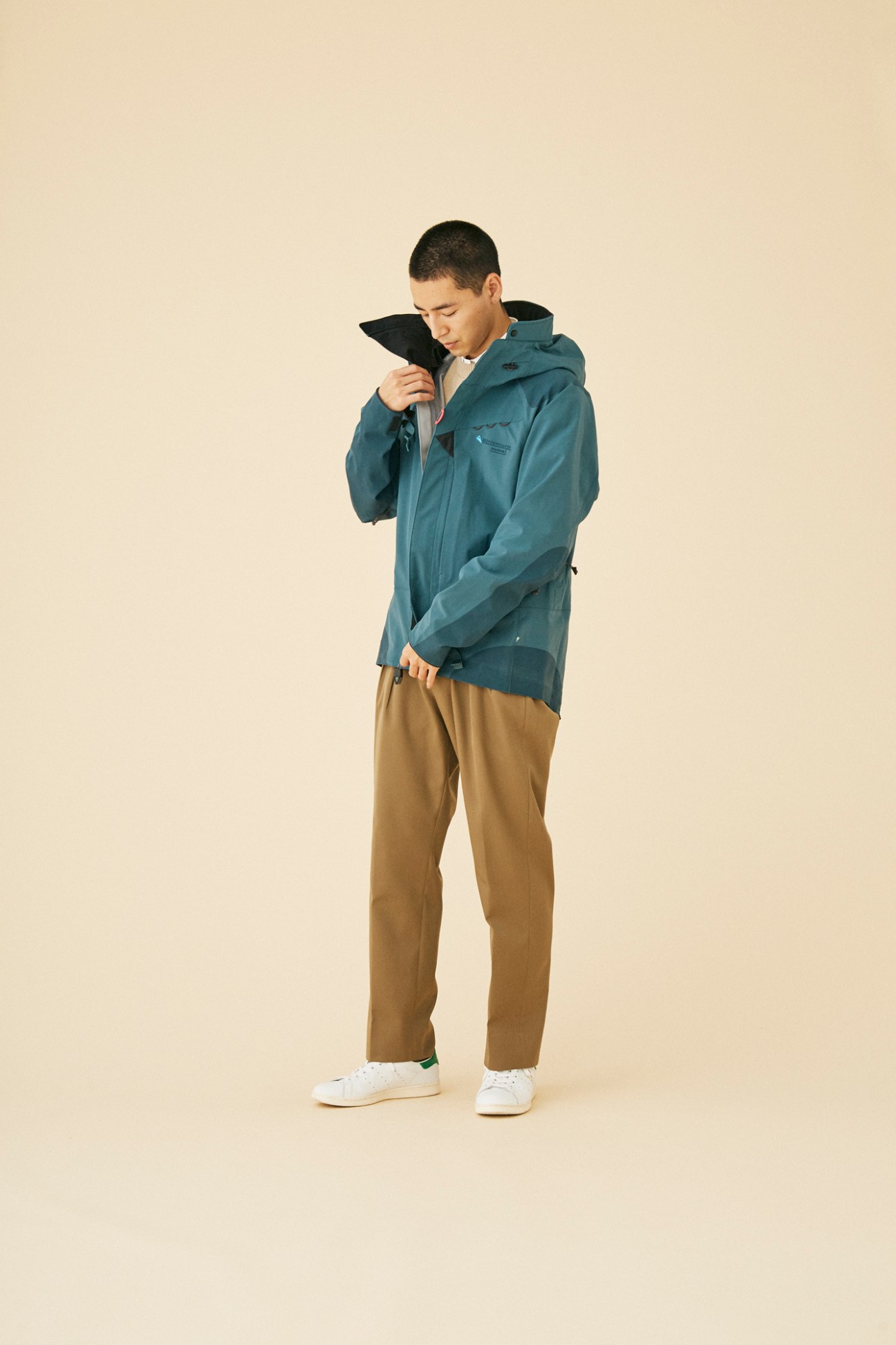 Man wearing teal technical jacket by Klättermusen with brown trousers and white sneakers against beige background