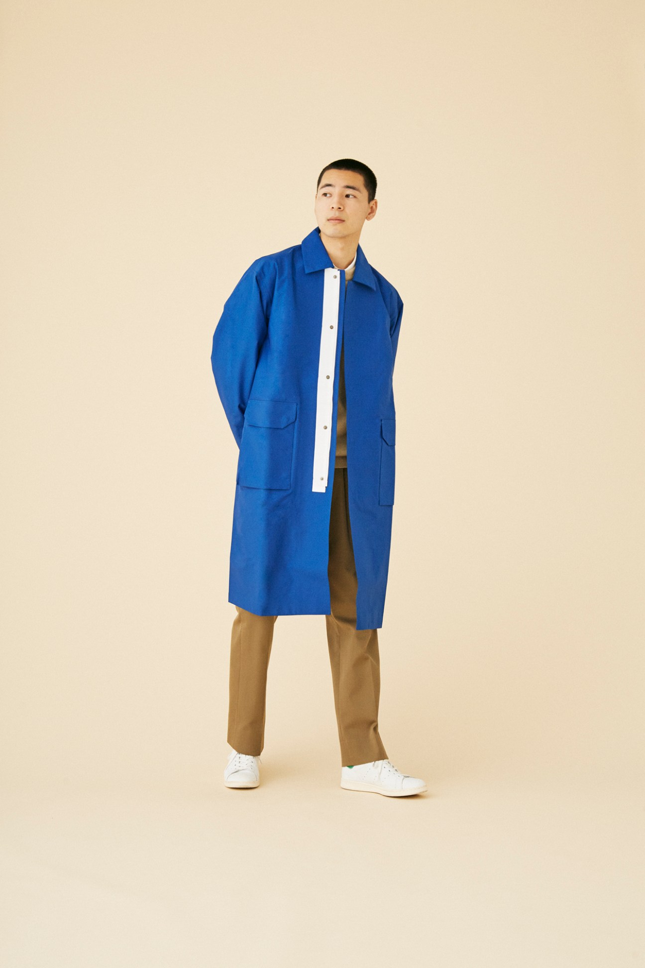 Man wearing royal blue long coat by F/CE with white shirt and brown trousers against neutral background
