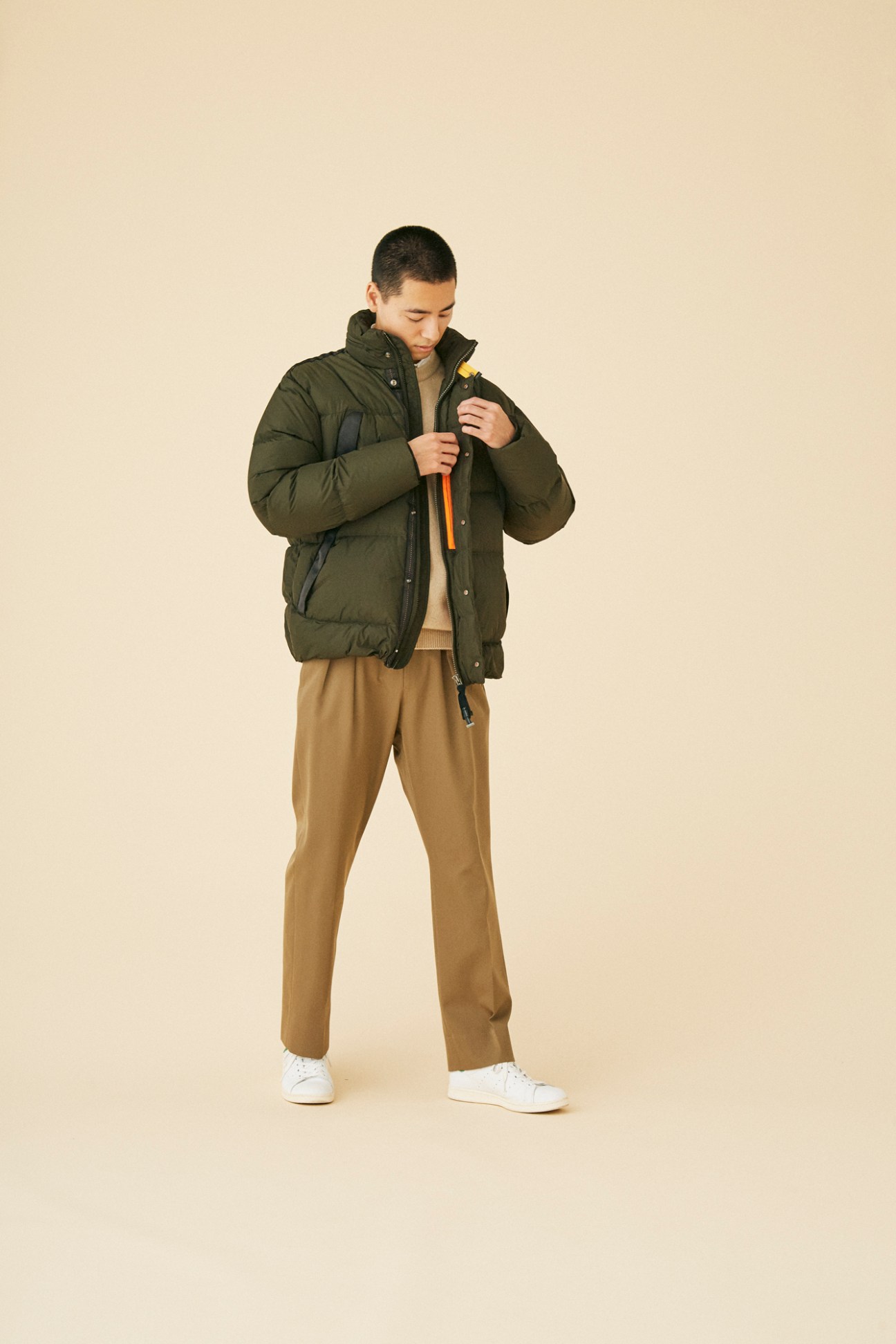 Man wearing olive green insulated jacket by Parajumpers with camel trousers and white sneakers in studio portrait