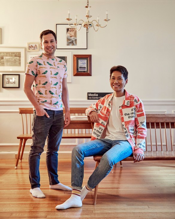 Two young men pose in an elegant interior space with mid-century furniture and framed artwork on the walls.
