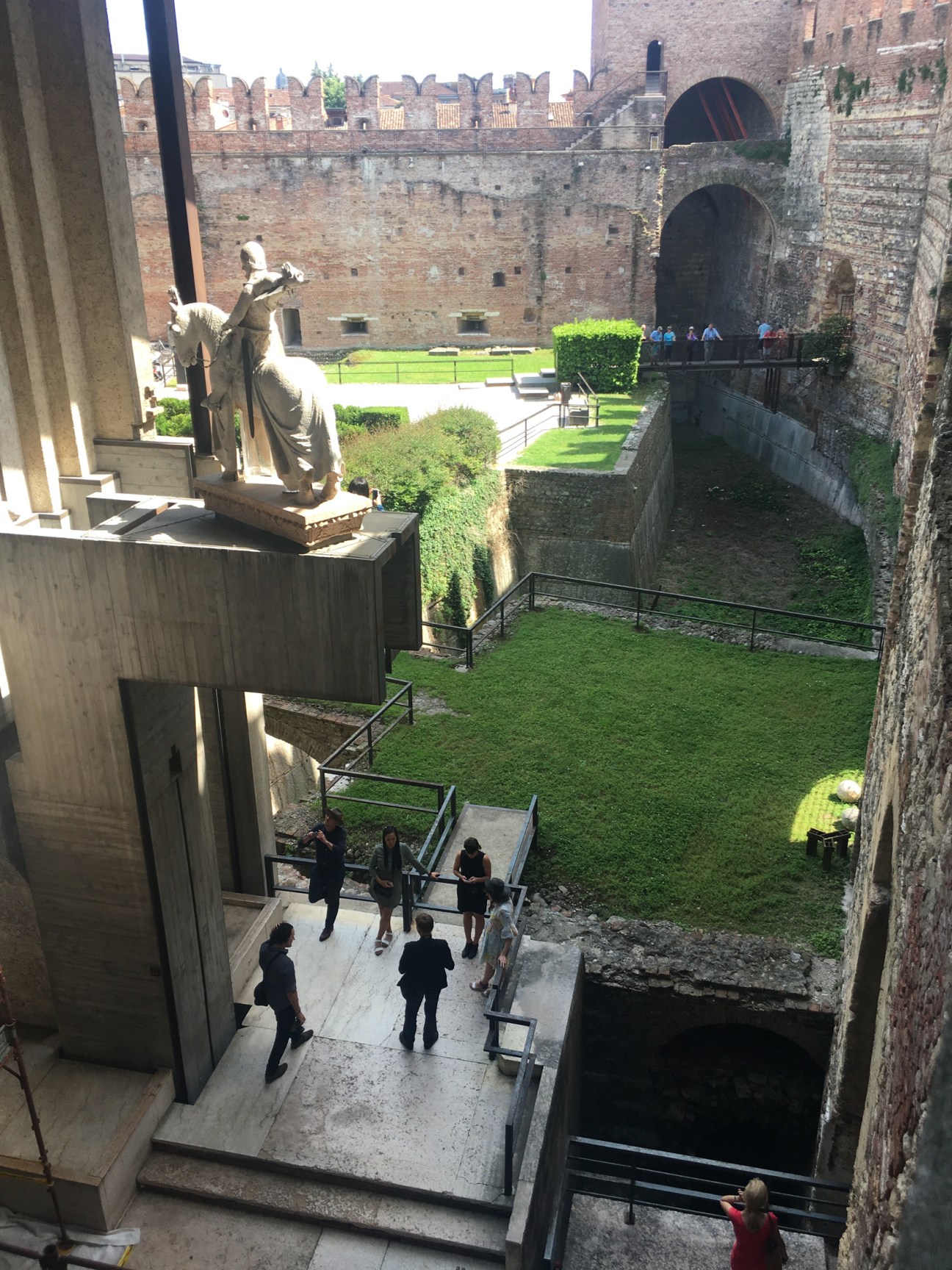 Stone courtyard with arched passages and grassy areas viewed from upper level with statue visible