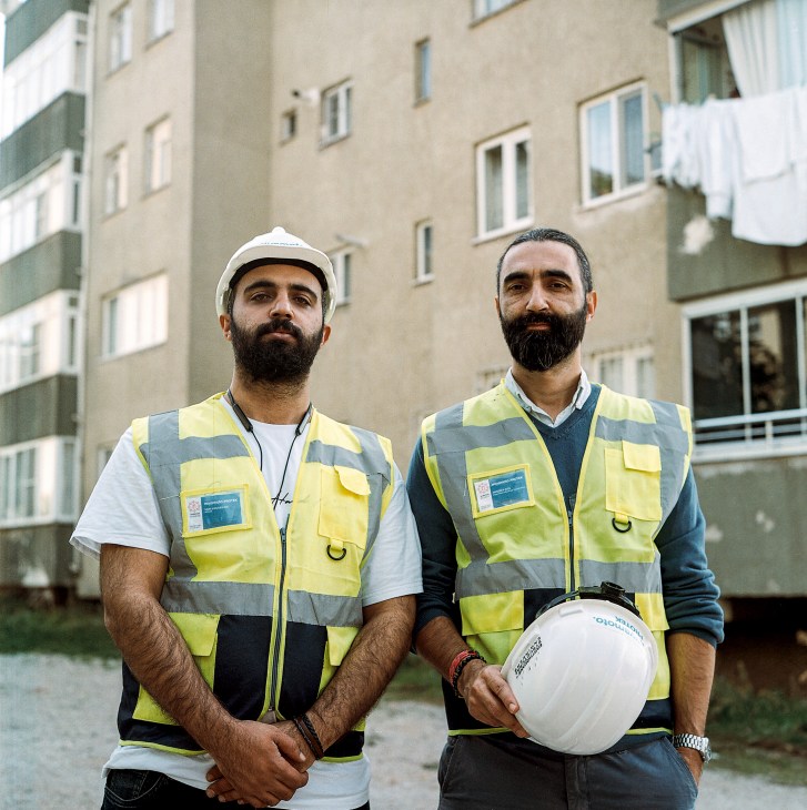 Two structural engineers in safety vests and hard hats stand in front of a damaged apartment building in Turkey.