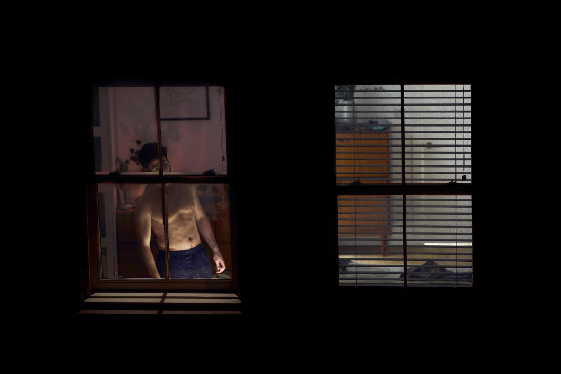 A shirtless man stands silhouetted against a window pane with warm golden light filtering through, captured by photographer Christopher Fernandez during London lockdown.