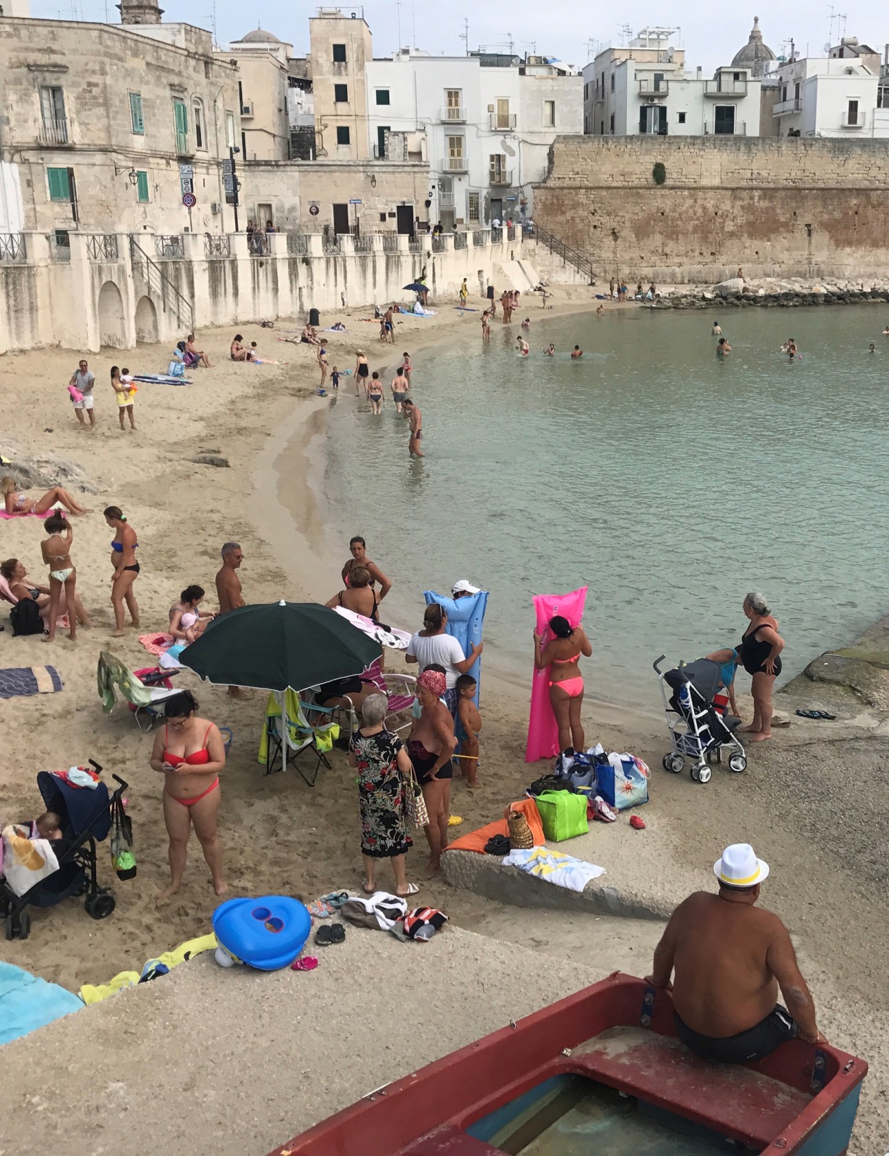 Beach scene with people swimming and sunbathing on sand near historic stone buildings and fortifications