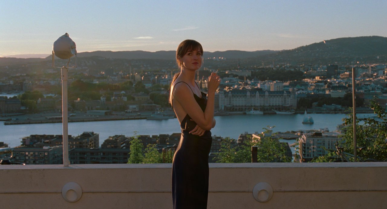 Woman in dark clothing making peace sign gesture overlooking city waterfront at dusk from rooftop or balcony