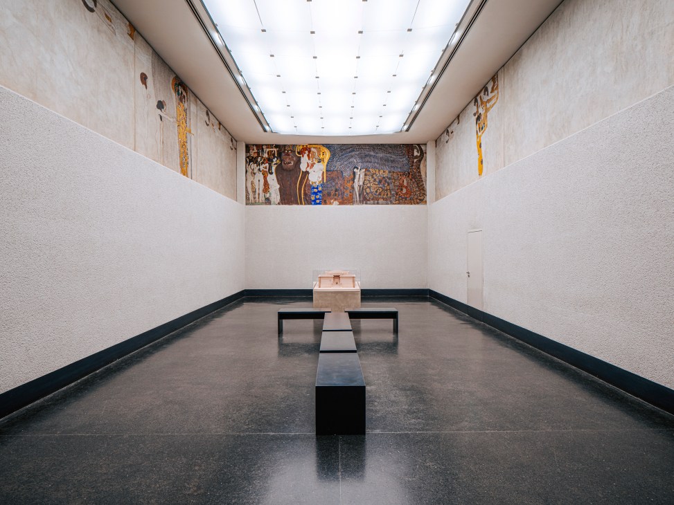 White museum interior with ornate ceiling frieze, viewing bench, and small display case
