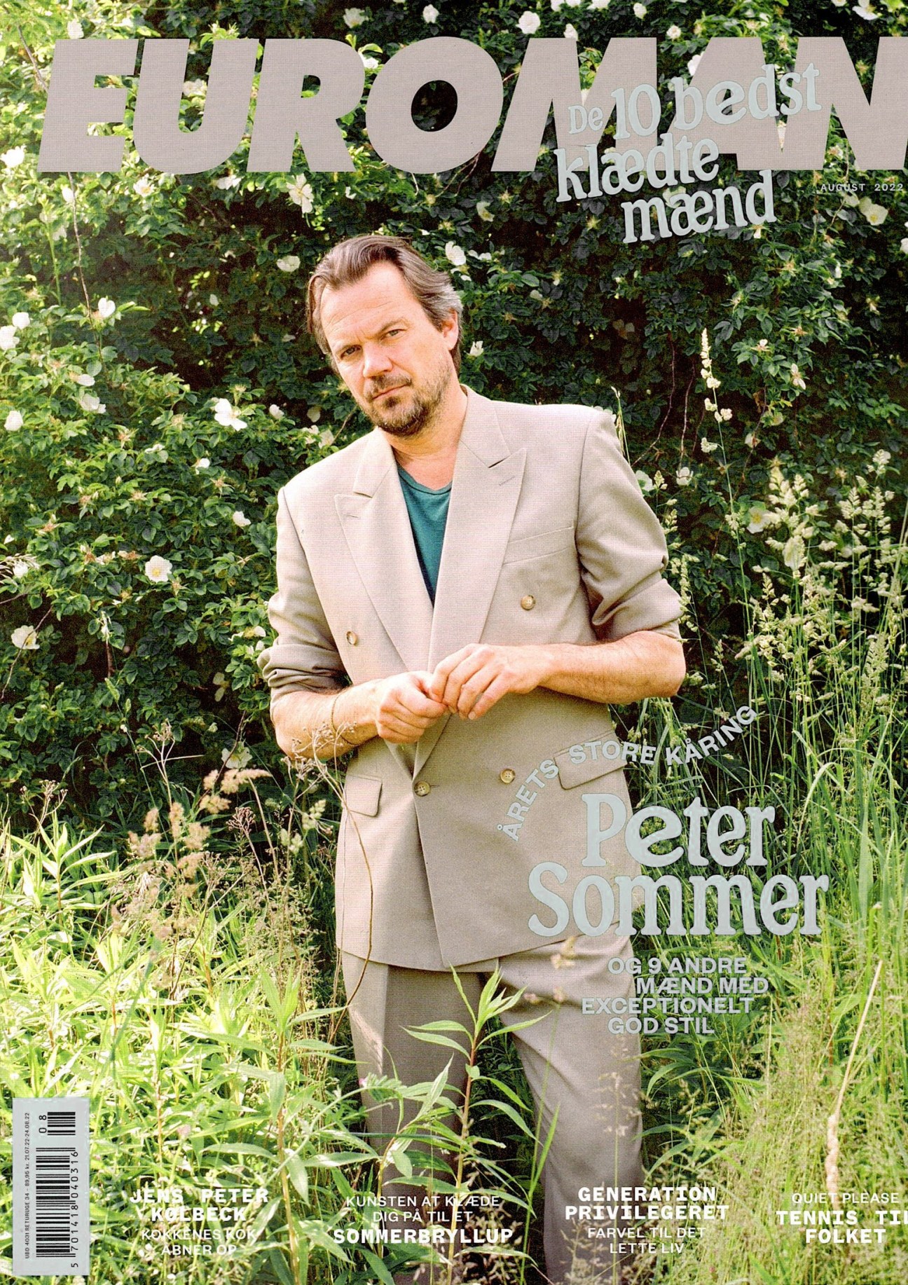 Man in beige double-breasted suit standing outdoors among green foliage and white flowers