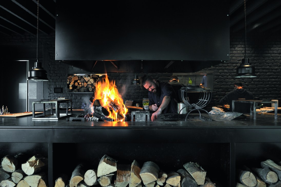 Chef working at wood-fired grill in professional kitchen with flames and firewood storage