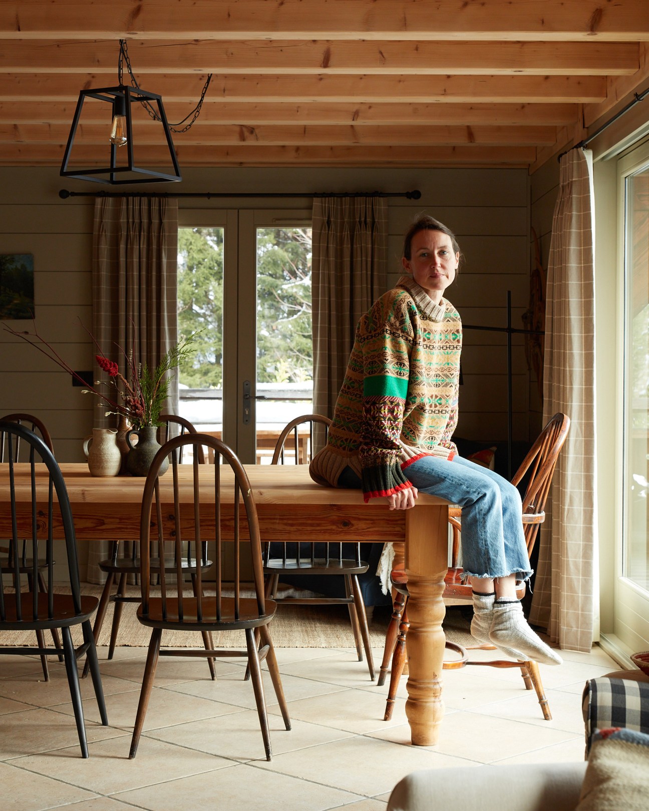 Designer Frances Stringer sitting on a wooden dining table in a rustic chalet interior wearing a patterned sweater