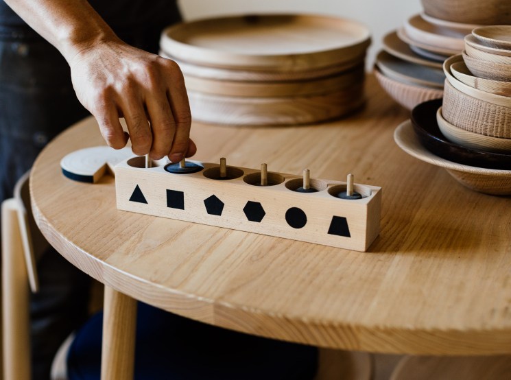 A hand places a piece into a wooden Montessori-style shape-sorting toy on a natural wood table, with stacked ceramics in the background.