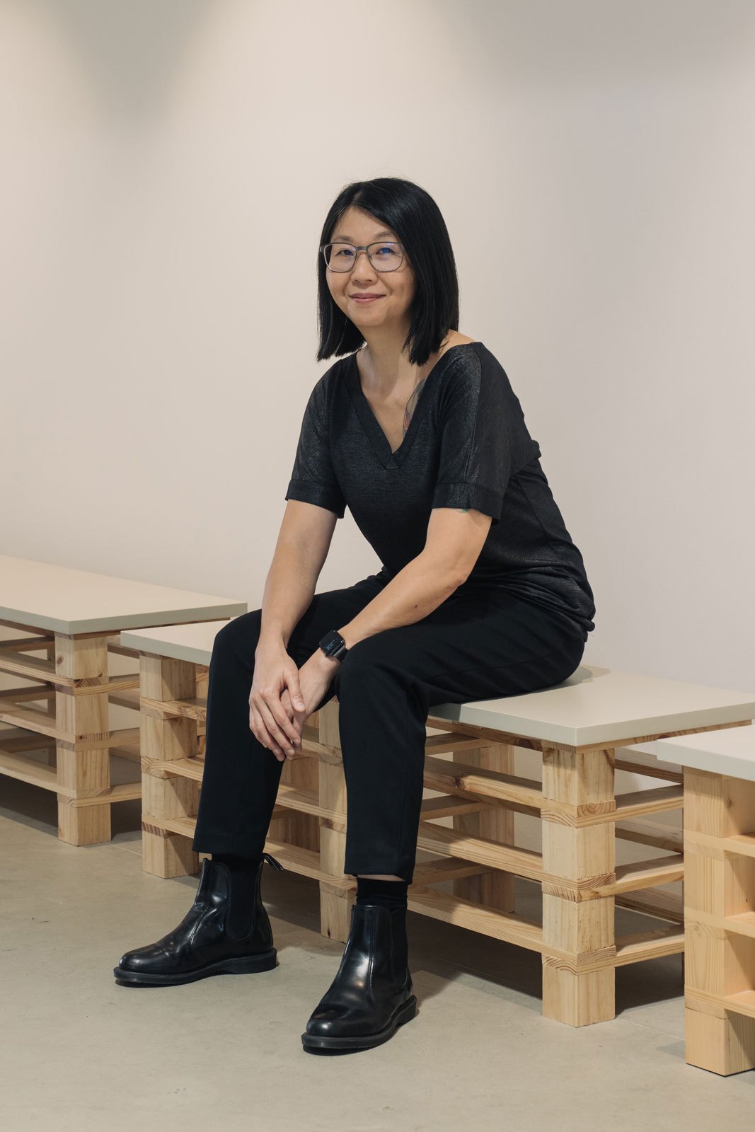 Woman sitting on wooden bench in gallery space wearing black clothing and glasses