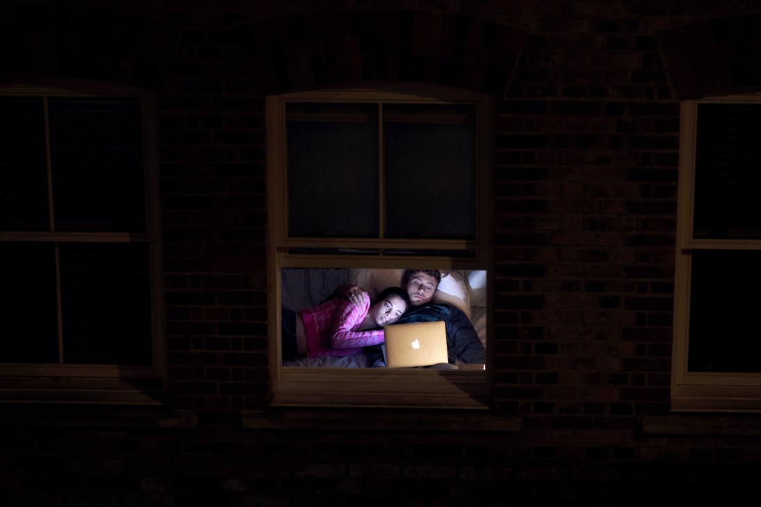 Two people in bed at night lit by a laptop screen, photographed through a window frame from outside.