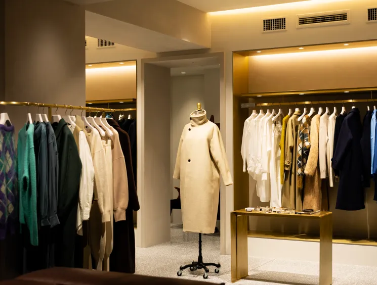 A warmly lit upscale fashion boutique with clothing racks and a cream coat displayed on a mannequin.