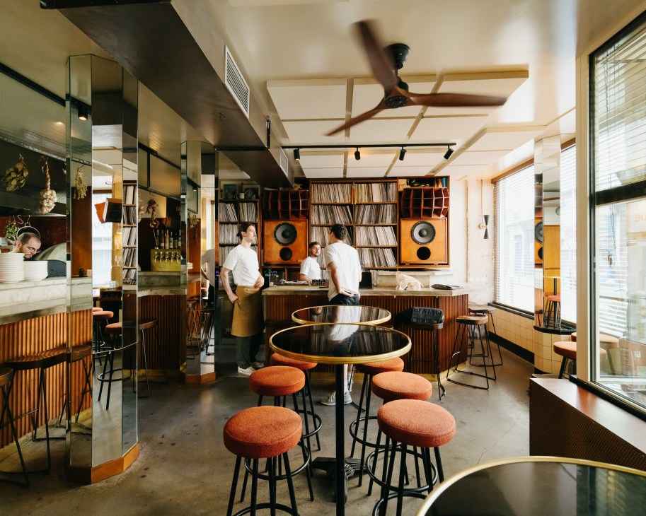 Modern bar interior with exposed beams, round tables with orange stools, vinyl records, and warm lighting