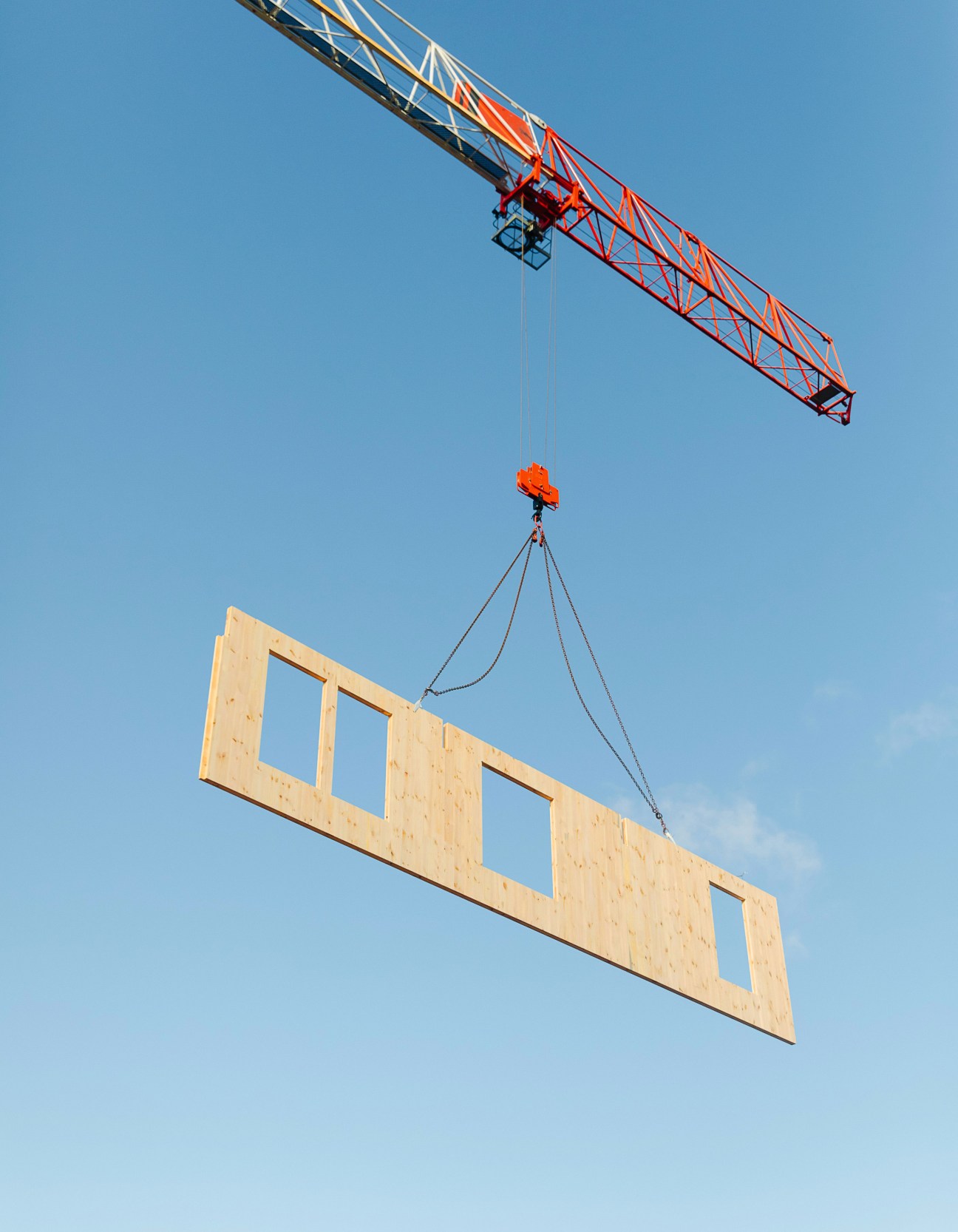A crane lifts a large prefabricated wooden panel against a clear blue sky at a construction site.