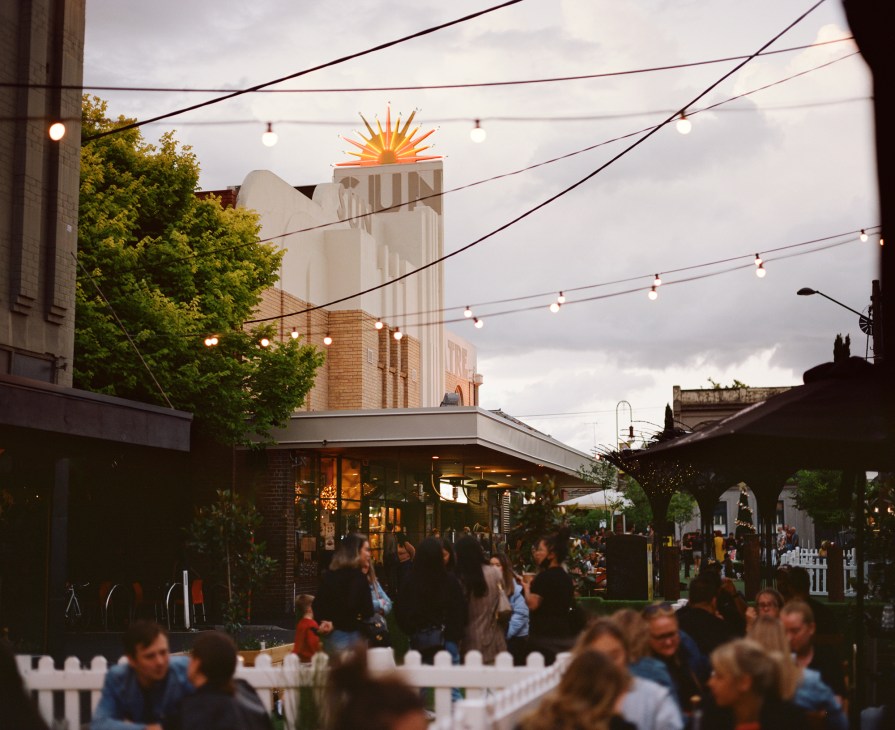 Evening scene with people gathering outdoors near buildings with string lights overhead and the Sun Theatre's illuminated sign