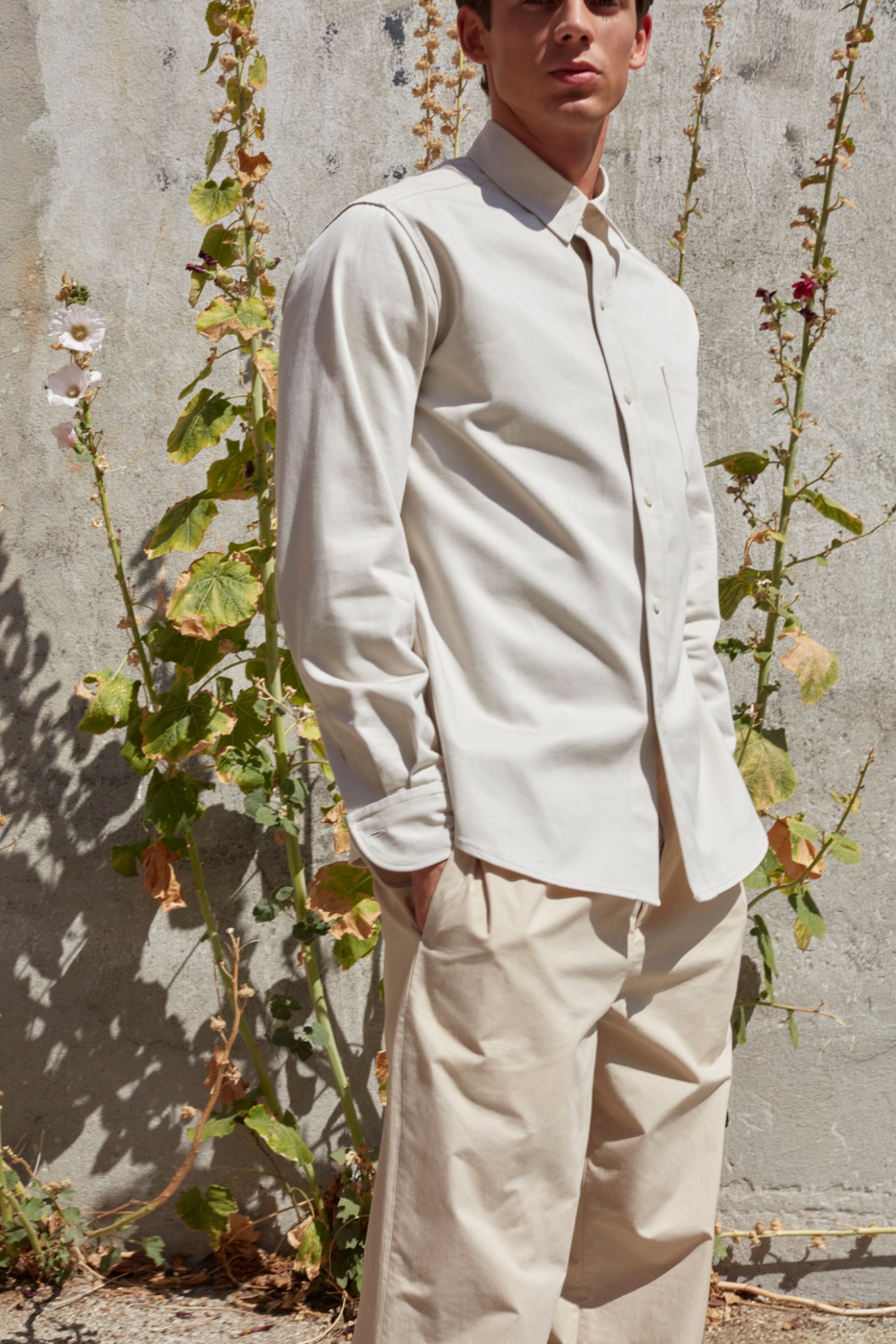 Man wearing beige button-up shirt and matching trousers standing against concrete wall with climbing vines