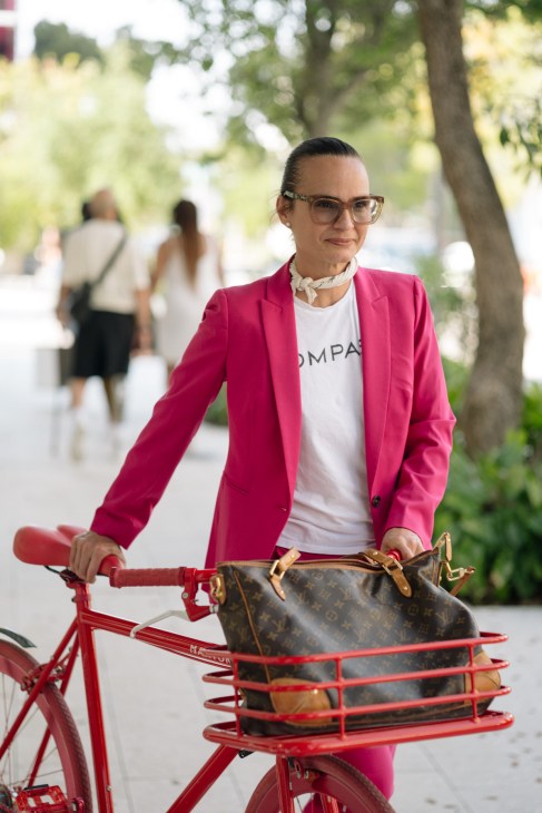 Woman in pink blazer with glasses on red bicycle with basket in tree-lined street.