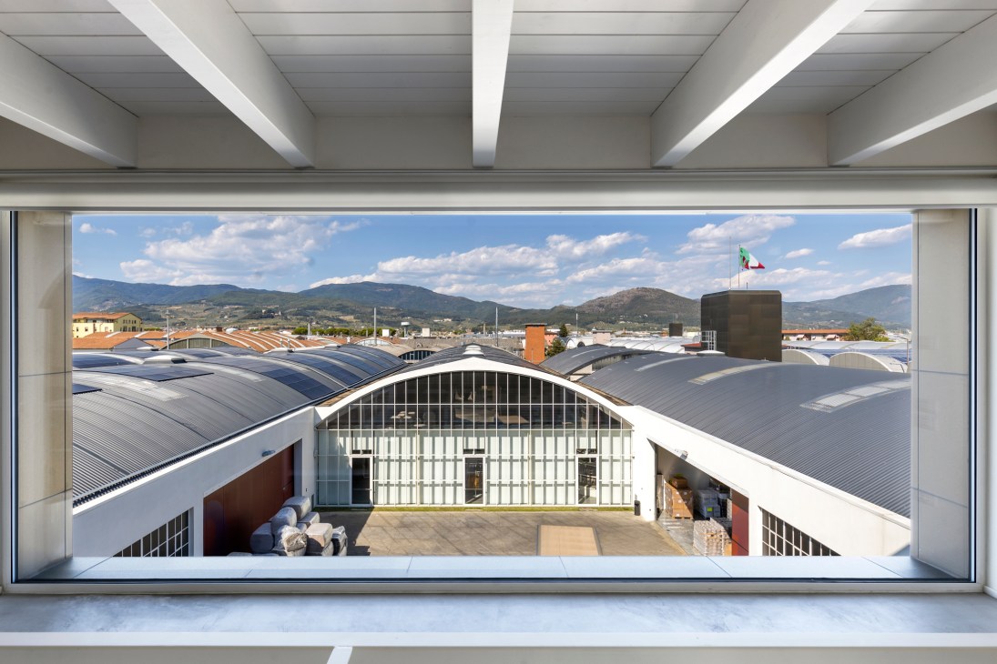 Modern industrial building with arched metal roofs viewed through large window frame with mountains in background