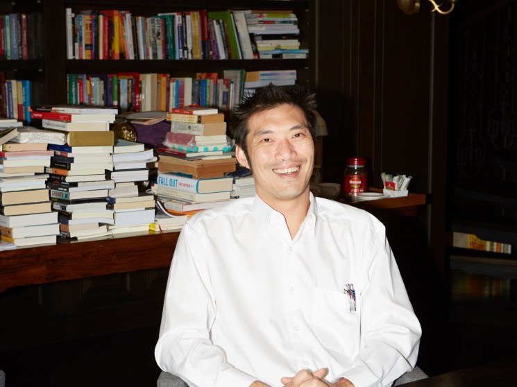 Smiling man in white uniform seated among stacks of books.