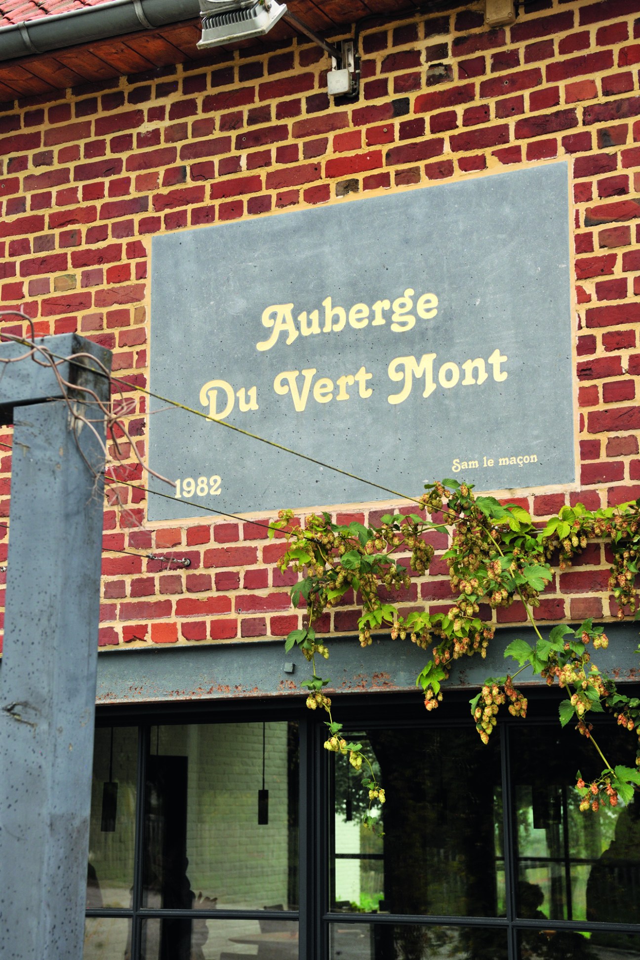 Stone sign reading "Auberge Du Vert Mont 1982" mounted on red brick wall with hop vines growing below