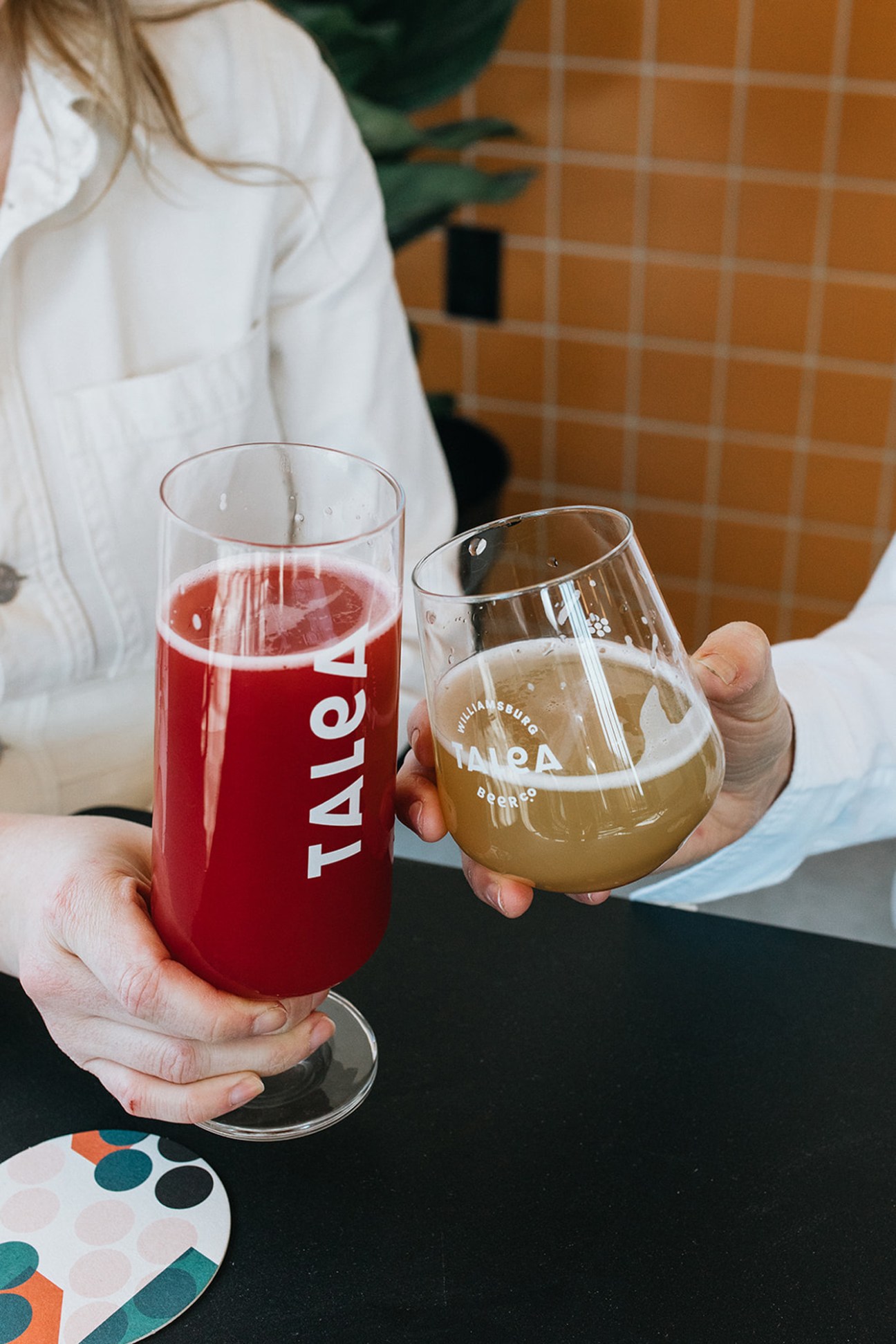 Two hands holding drinks at Talea brewery - a tall glass with red beverage and a stemmed glass with light beer