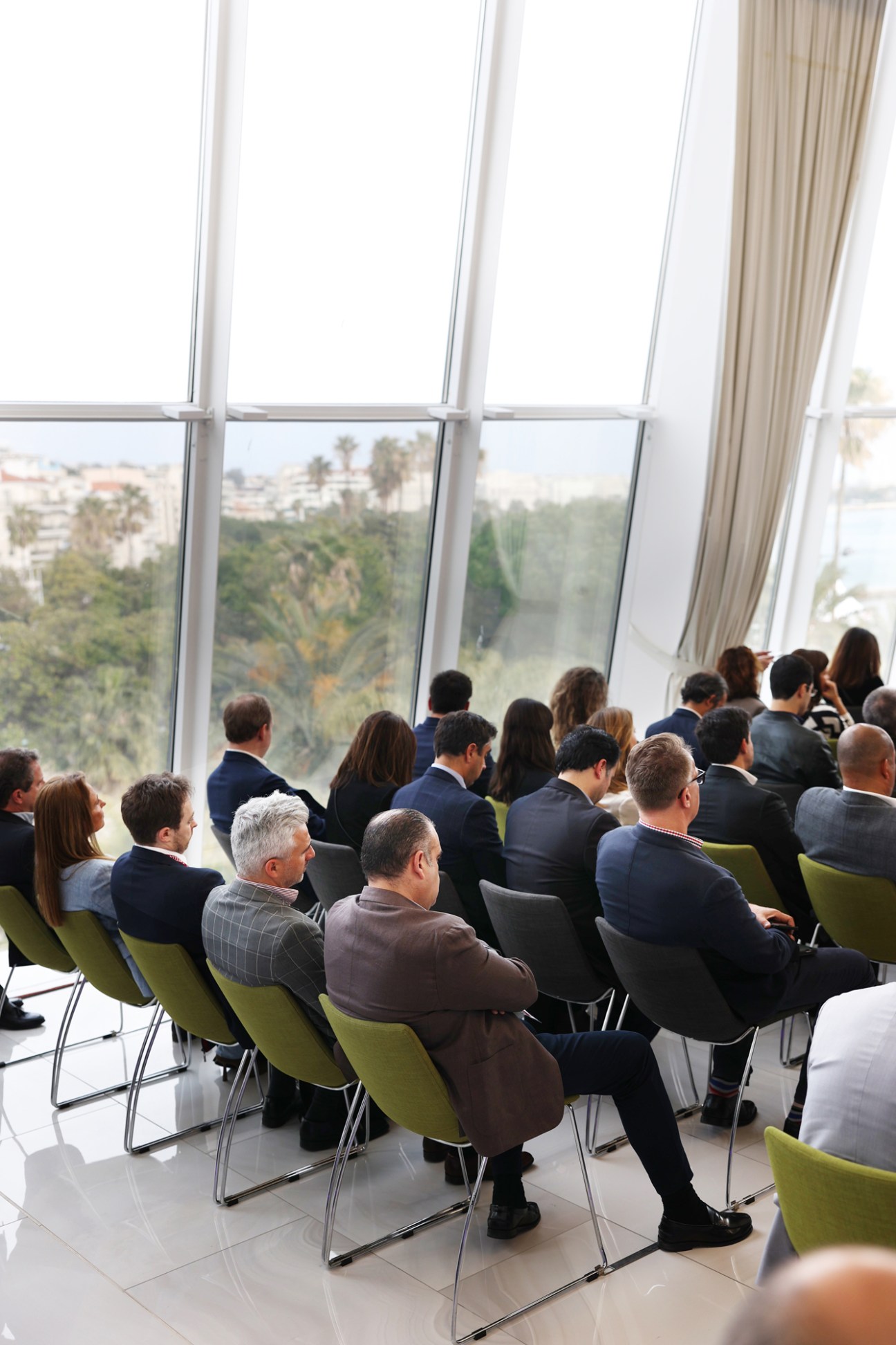Business professionals in suits seated in green chairs at conference presentation with large windows