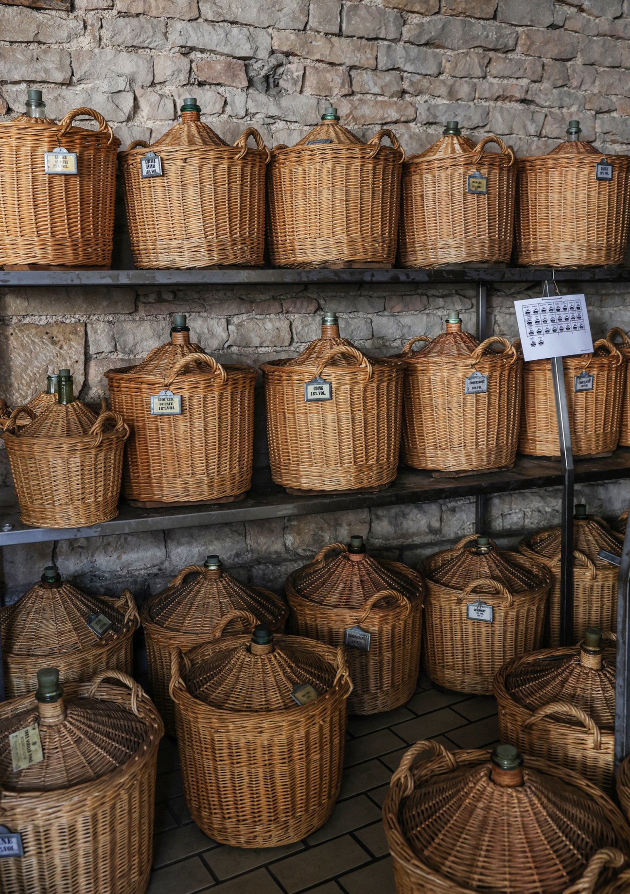 Traditional wicker demijohns with glass bottles lined on wooden shelves against stone wall in Burgundy wine cellar