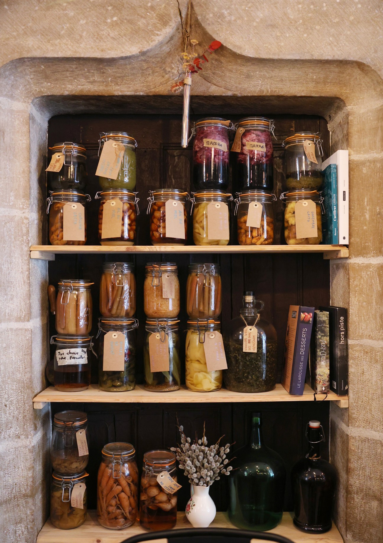 Glass jars with hinged lids containing pickled vegetables displayed on wooden shelves in a stone alcove at Betterave
