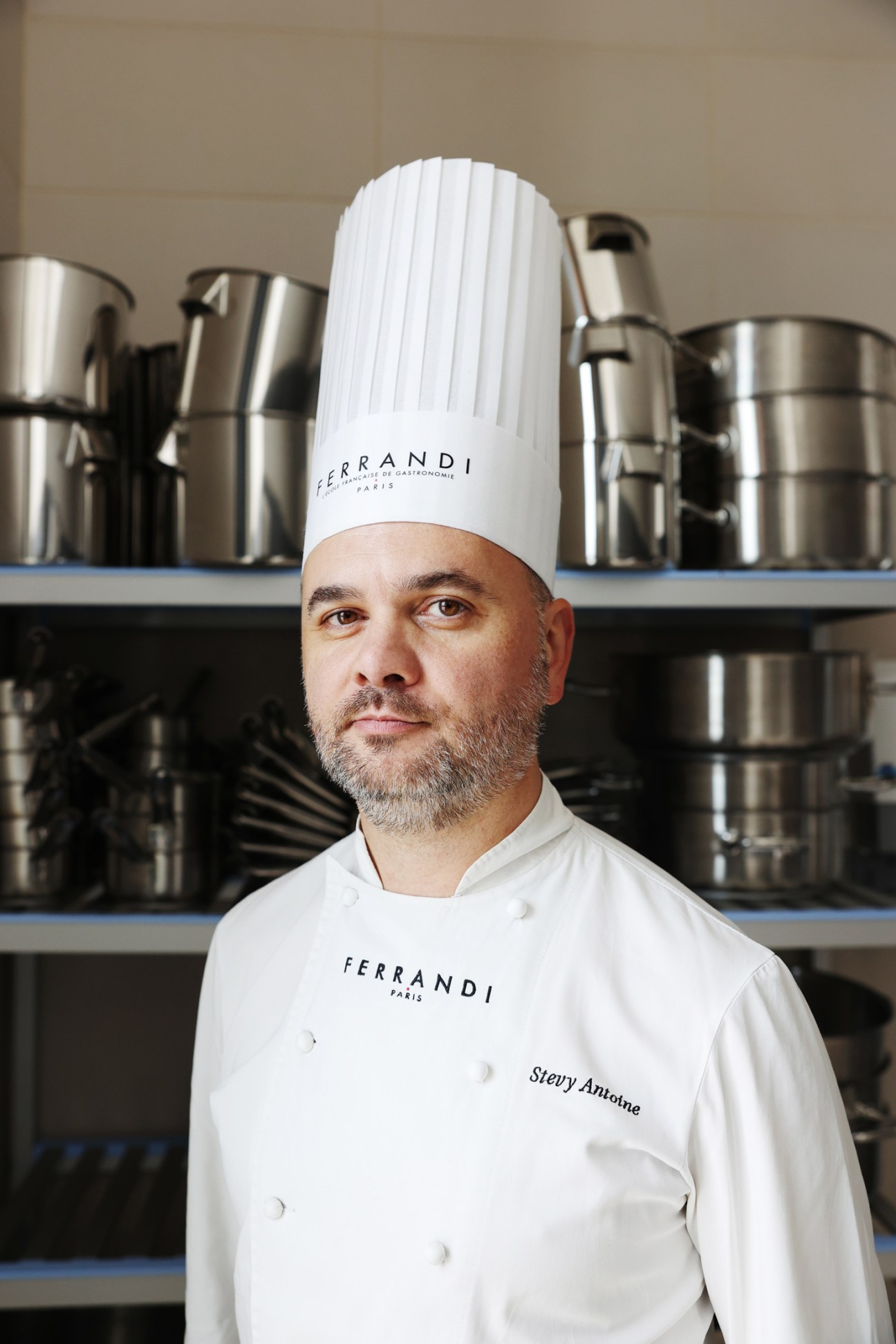 Stévy Antoine in white chef's uniform and tall toque standing in professional kitchen with stainless steel pots on shelves