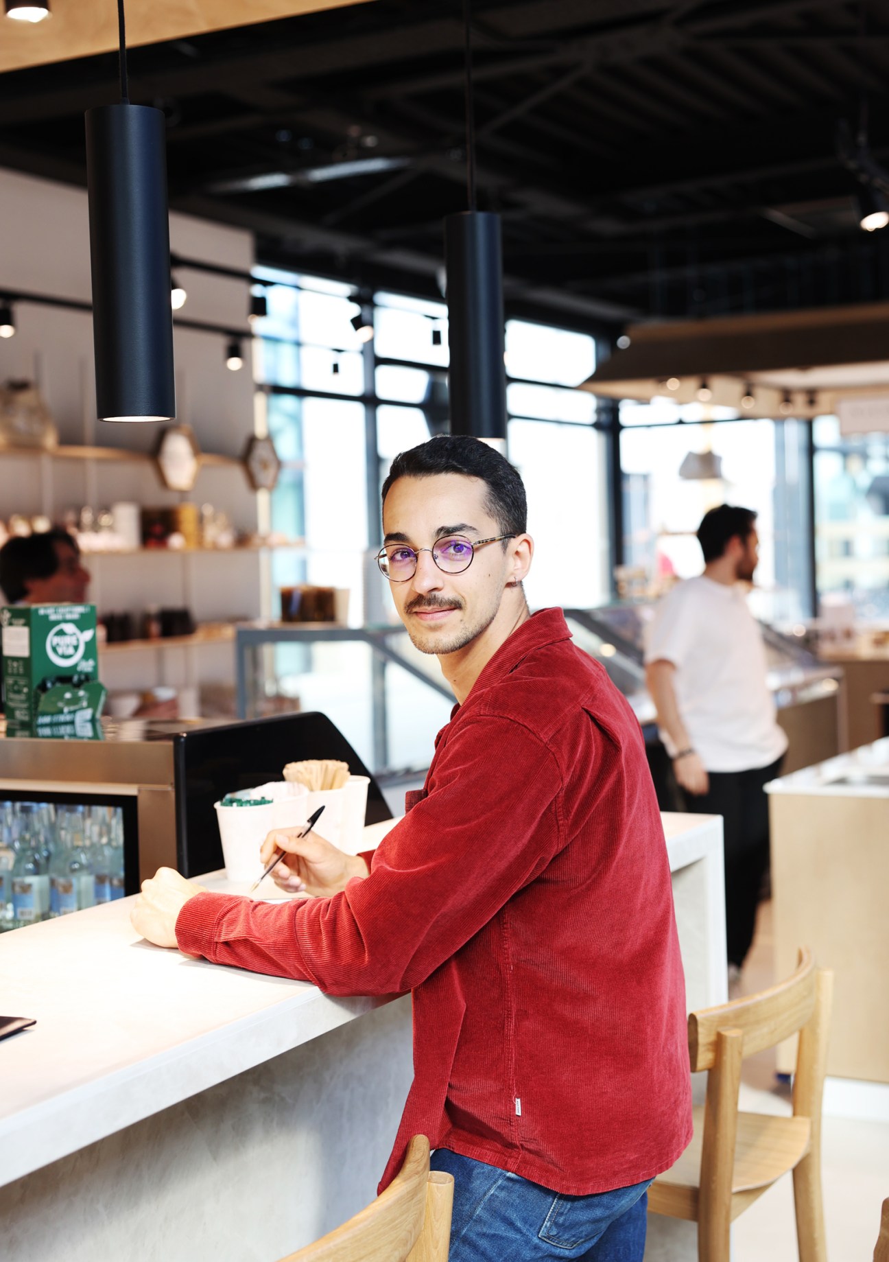 Man in red sweater and glasses writing at modern cafe counter with industrial lighting and large windows