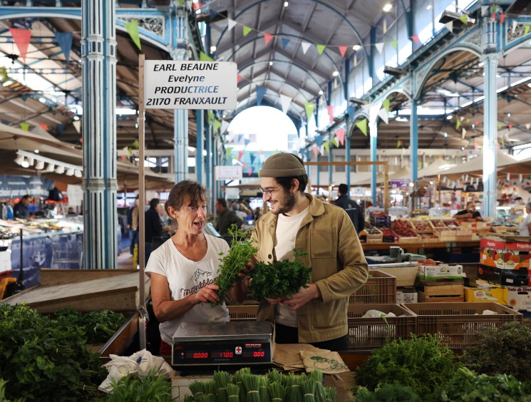 Chef Angelo Ferrigno with a woman vendor smiling while holding leafy greens at a produce stand in Dijon's historic covered market