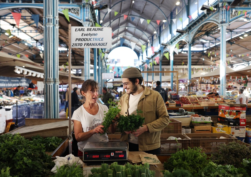 Chef Angelo Ferrigno with a woman vendor smiling while holding leafy greens at a produce stand in Dijon's historic covered market