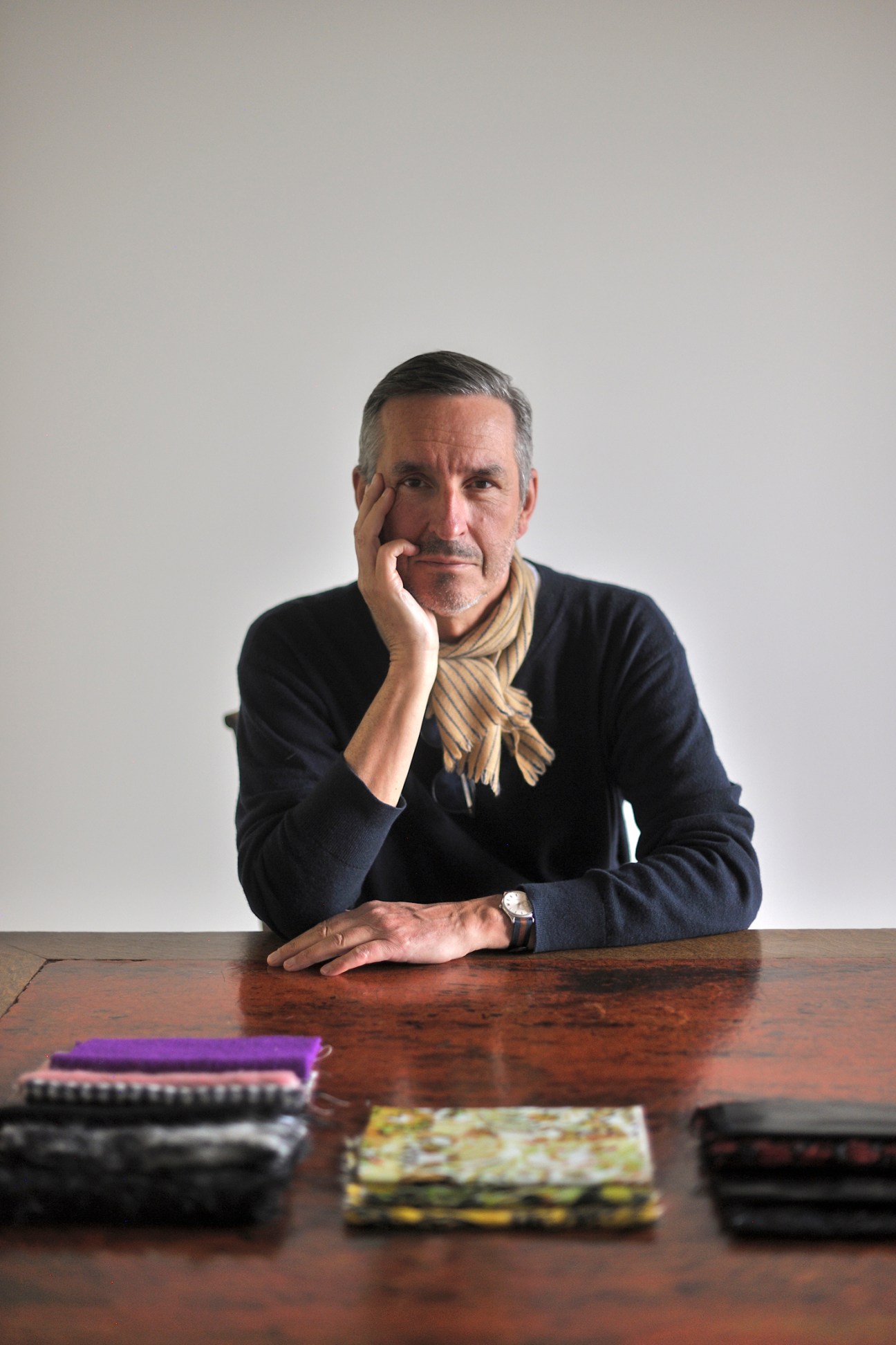 Fashion designer Dries Van Noten sitting at wooden desk with hand on chin in contemplative pose