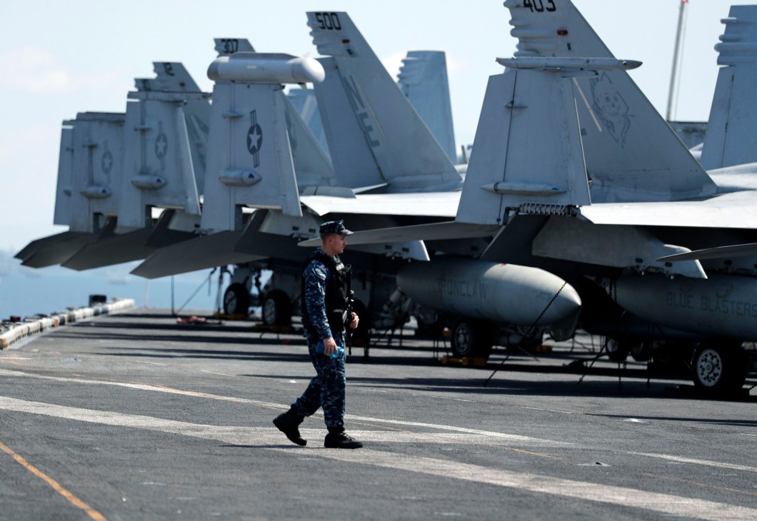 Naval officer in blue camouflage uniform walking across aircraft carrier flight deck with fighter jets