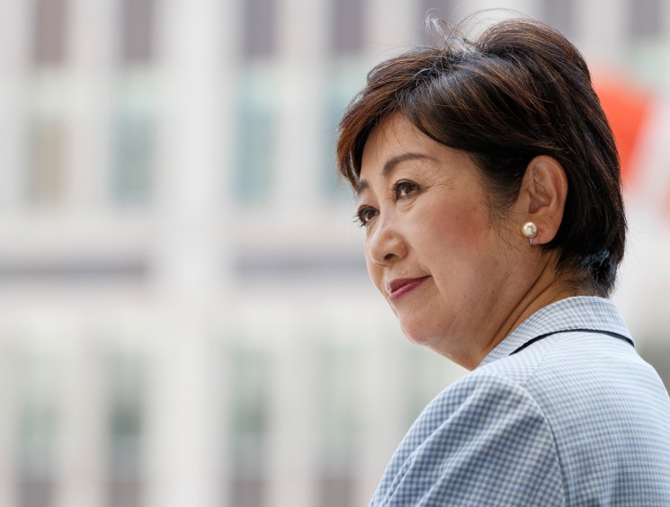 A side profile portrait of a woman in a light blazer with pearl earrings, looking into the distance against a soft urban backdrop.