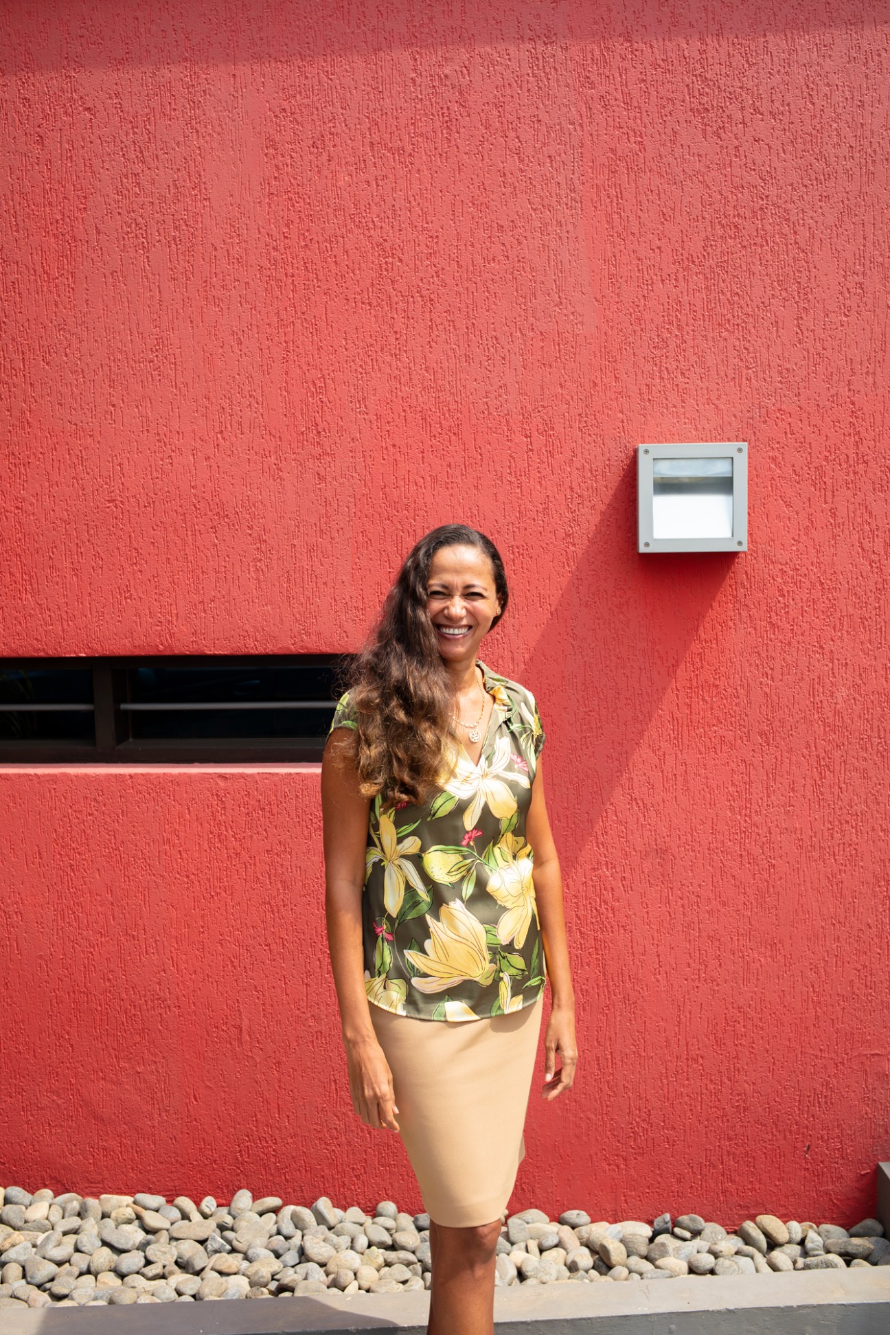 Rosemary Orthner stands smiling against a red textured wall wearing a tropical print top and beige skirt