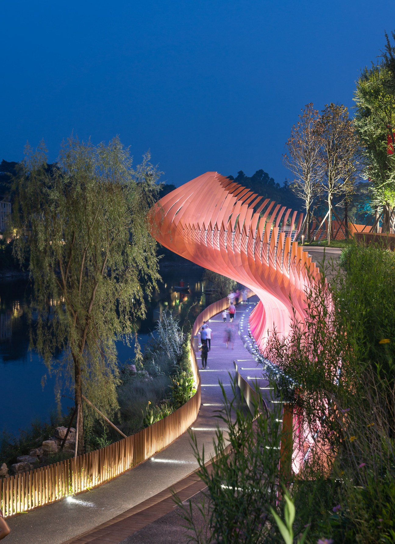 Illuminated curved pedestrian bridge with orange sculptural canopy crossing water at dusk in Zigong Dongxingsi Riverfront Park
