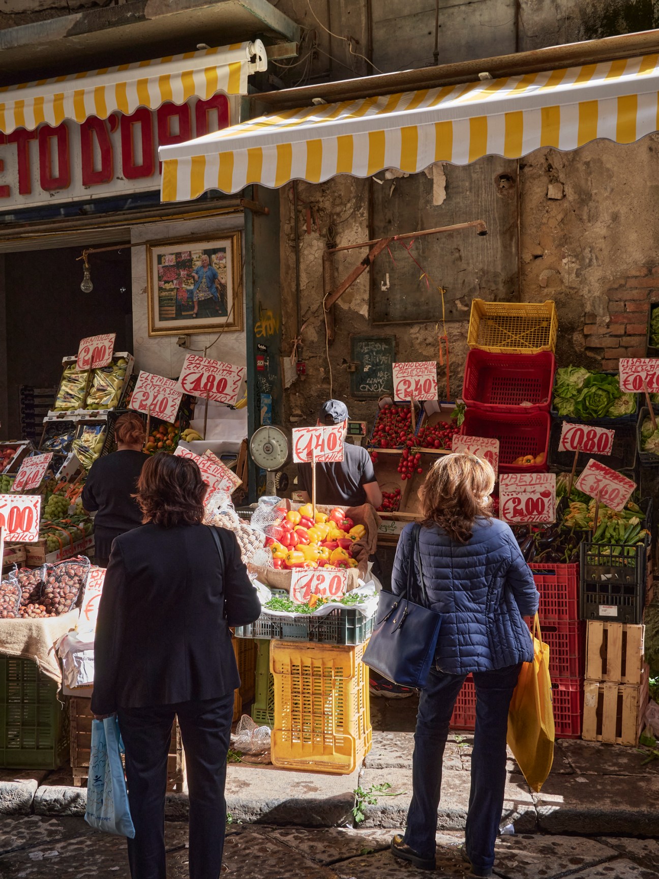 Two women shopping at an Italian food market stall with fresh produce and price signs
