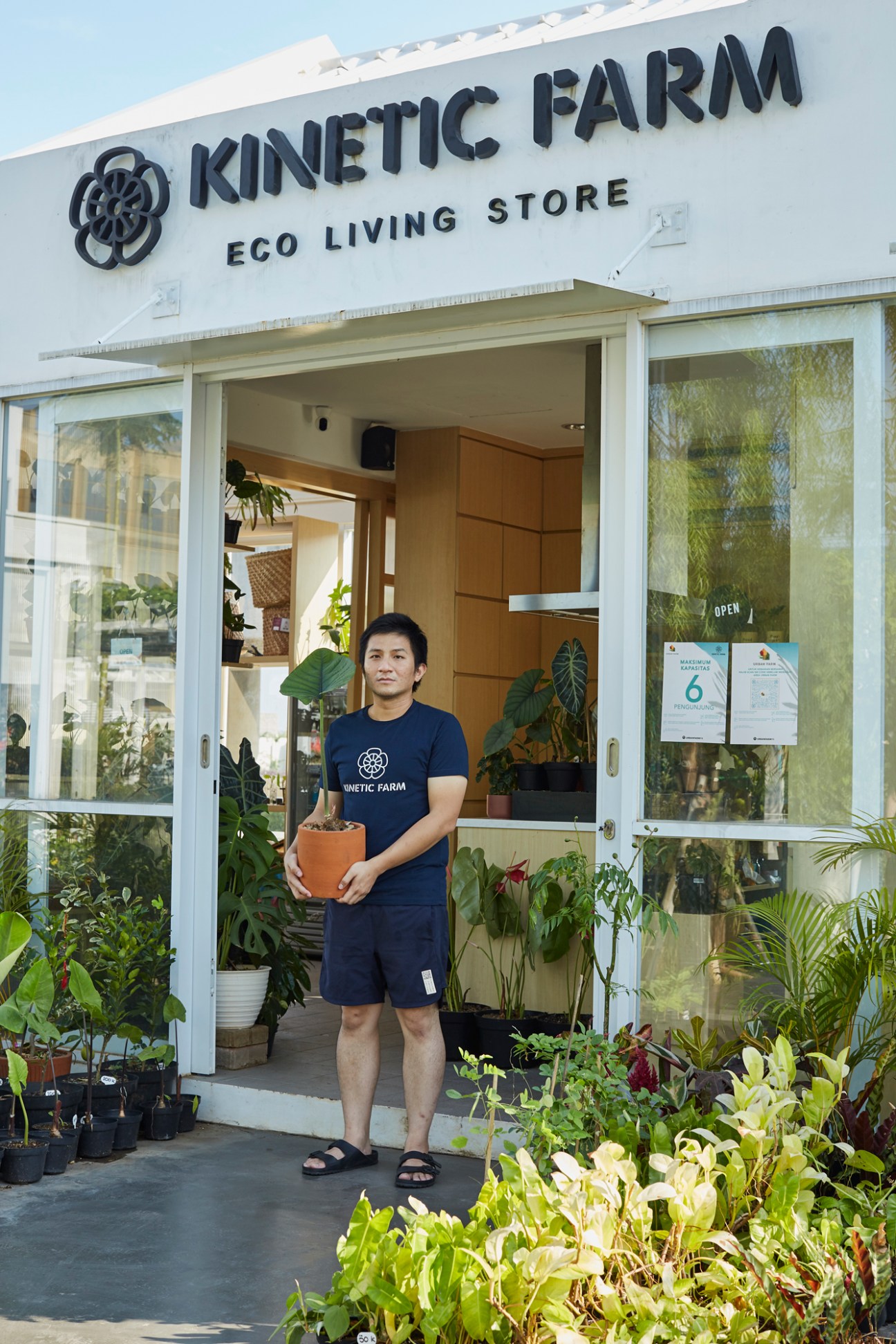 Man in Kinetic Farm navy t-shirt holding potted plant outside eco living store entrance in Indonesia