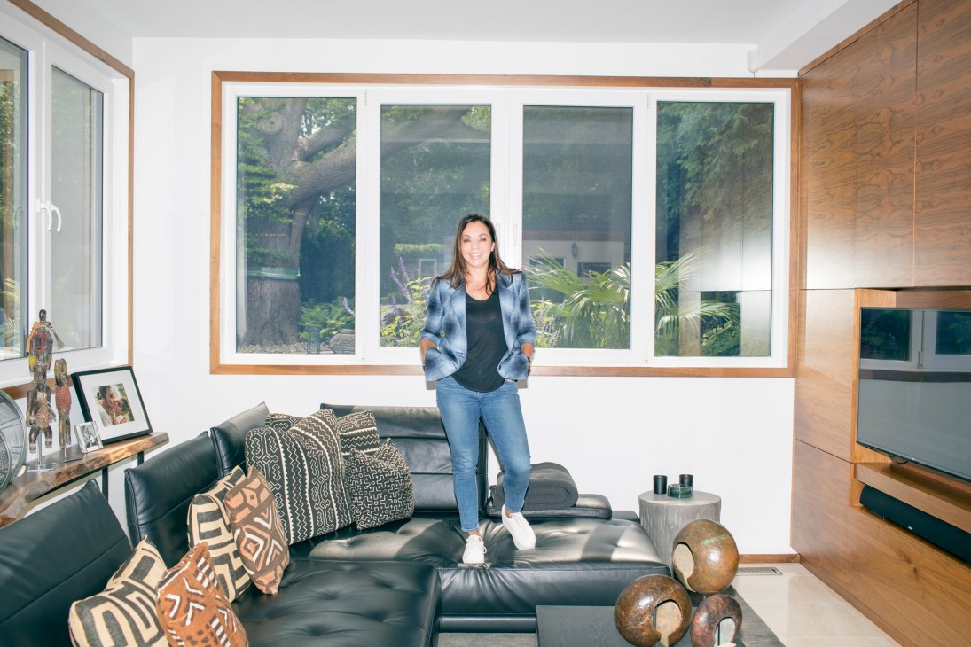 Woman standing in modern living room with large windows overlooking garden, black leather sofa and wooden furnishings.