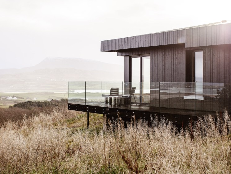 A dark timber modernist house with a glass-railed deck overlooking a vast misty highland landscape.
