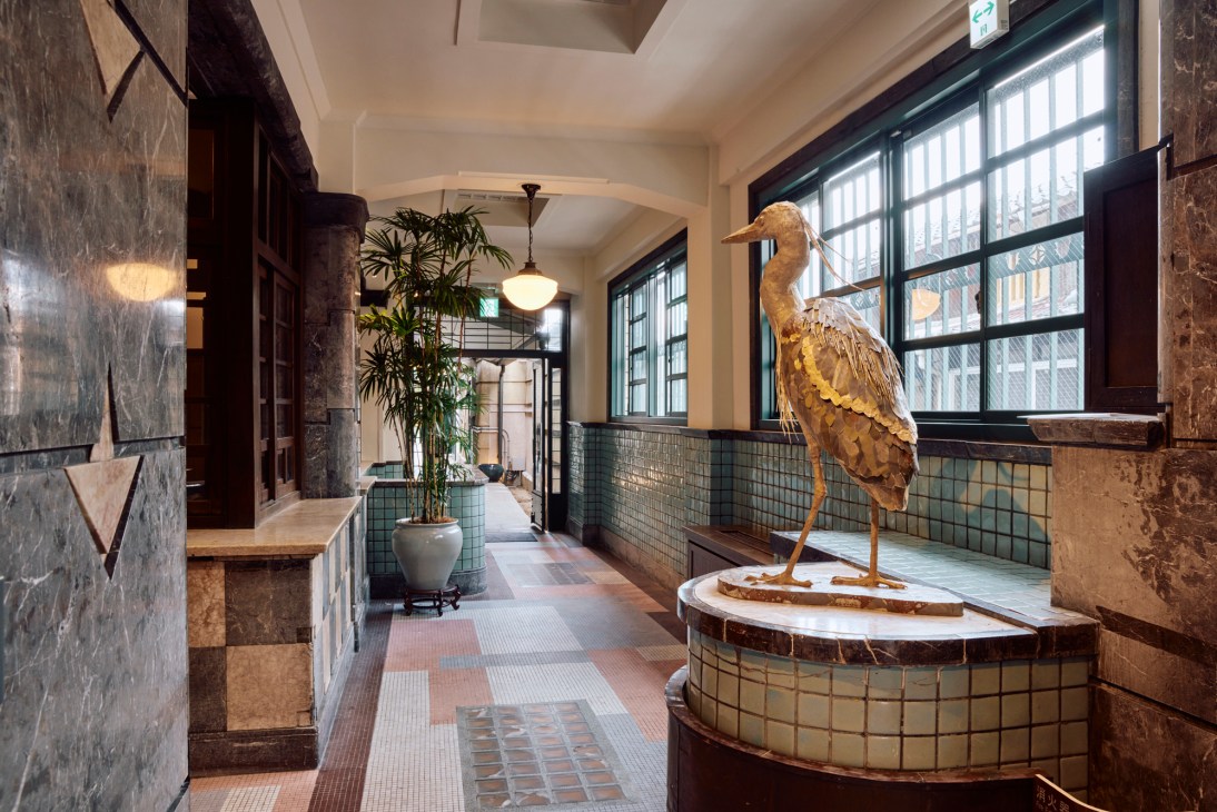 Decorative bronze stork sculpture on tiled pedestal in hotel corridor with large windows