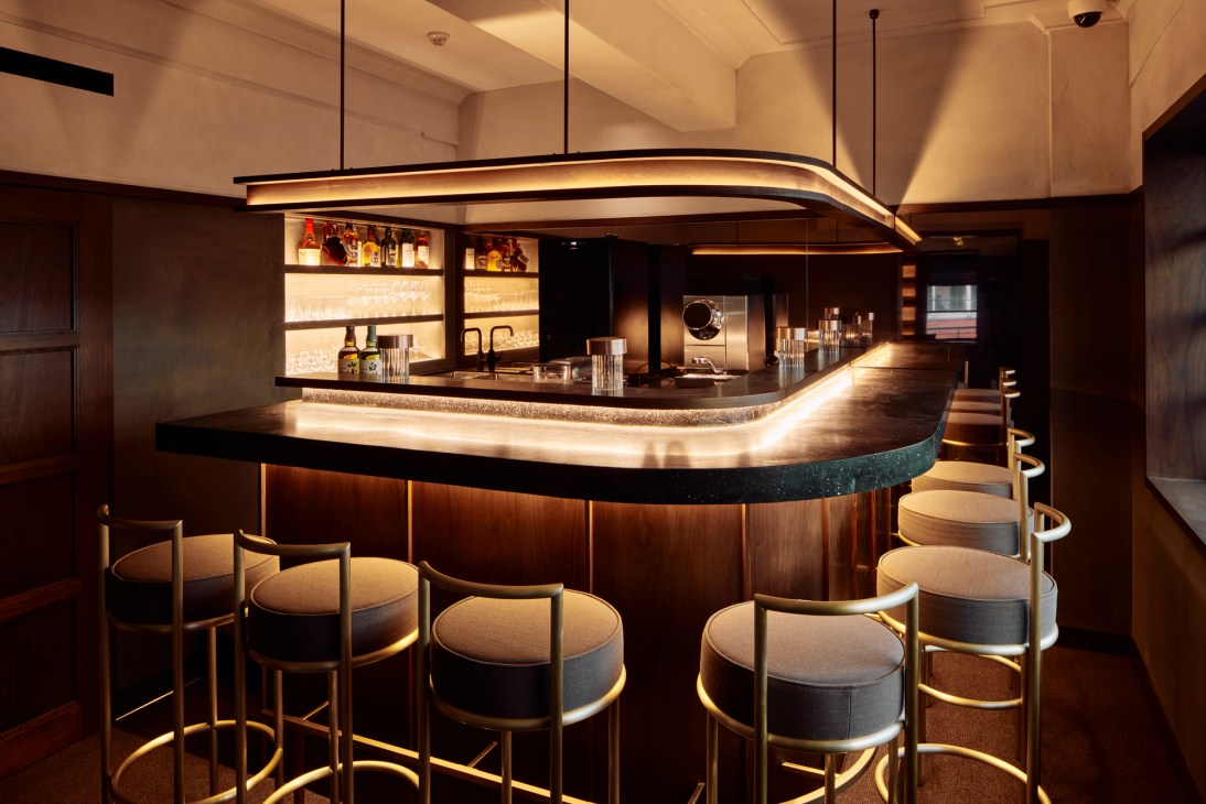 Modern hotel bar with curved wooden counter, illuminated shelving, and upholstered bar stools in warm lighting