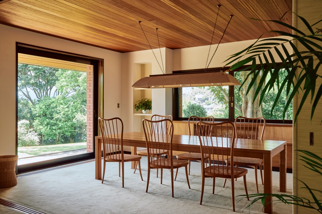 A dining room with wooden table, chairs, and pendant light, surrounded by large windows overlooking lush green trees