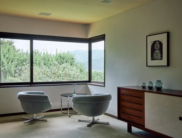 Modern interior corner with gray swivel chairs, glass side table and wooden credenza near large windows